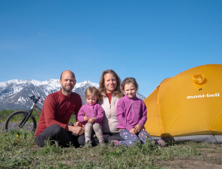 パッシュファミリーが晴れた空の元、雪に覆われた山々をバックに、テントの横で座っている写真